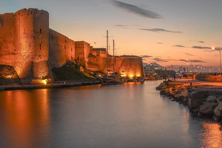 Stone fortress walls illuminated at sunset beside a calm harbor with a docked sailboat.