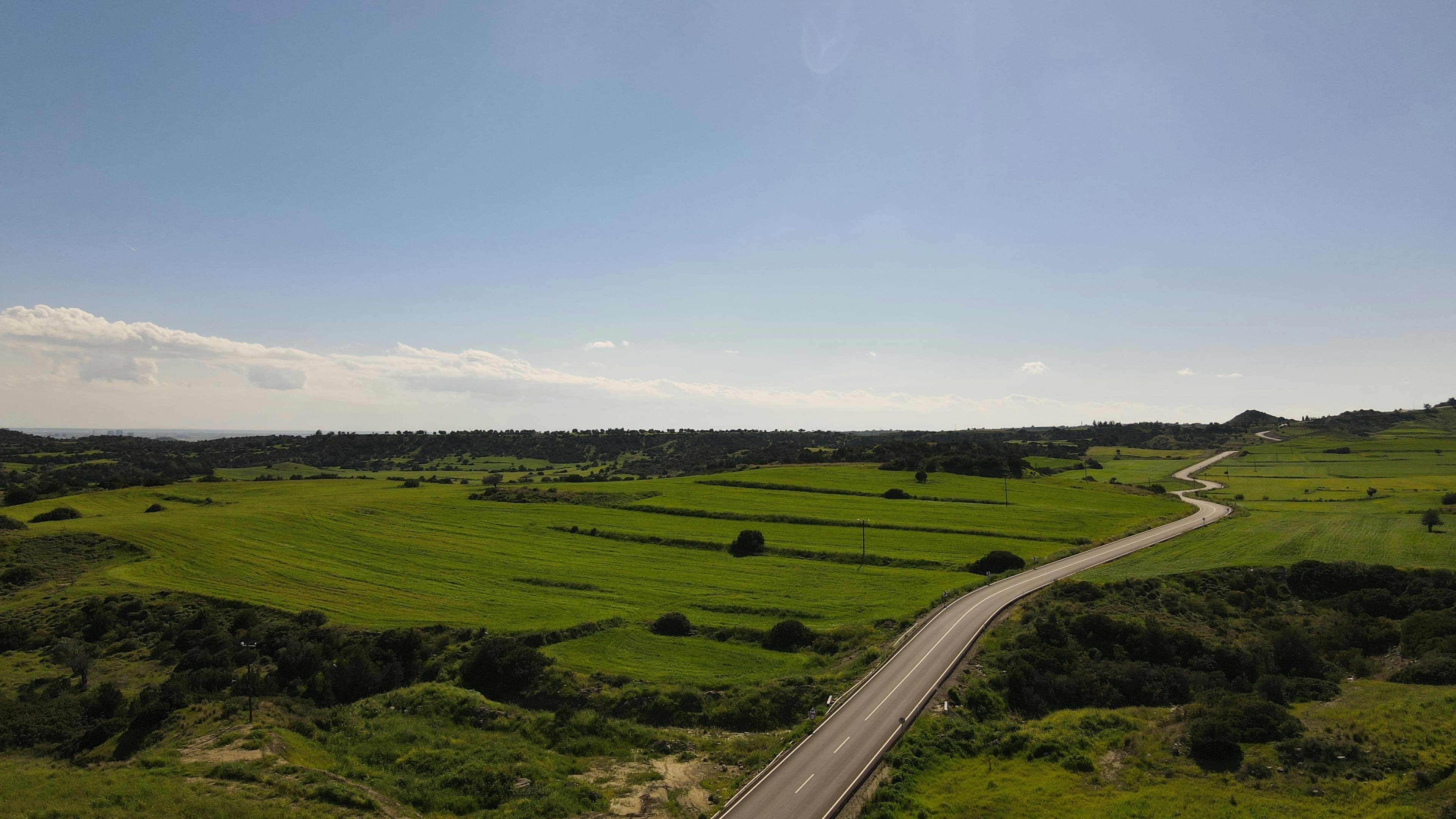 Winding road cutting through lush green fields and rolling hills under a clear blue sky.