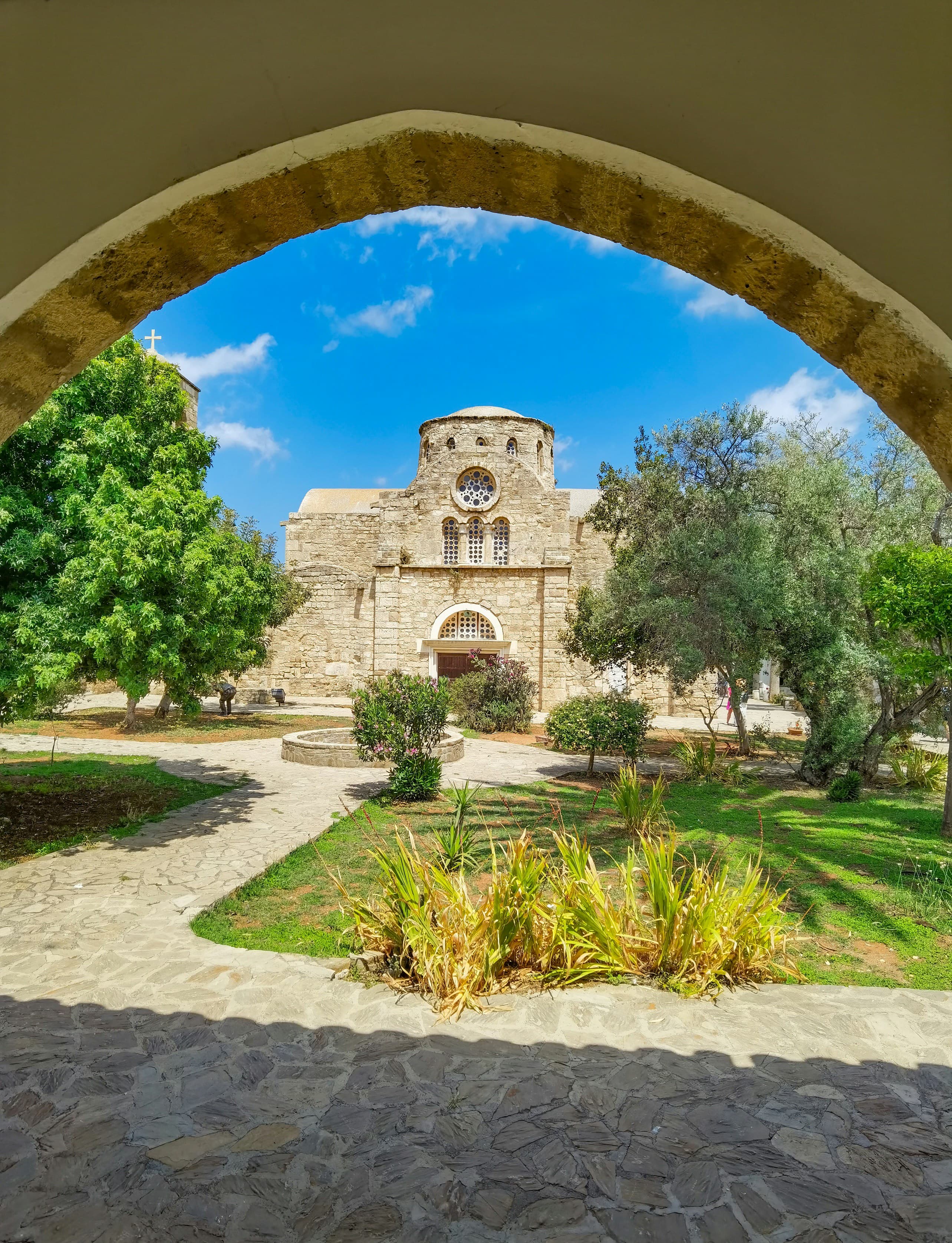 Ancient stone church with a dome framed by an archway in a lush courtyard garden.
